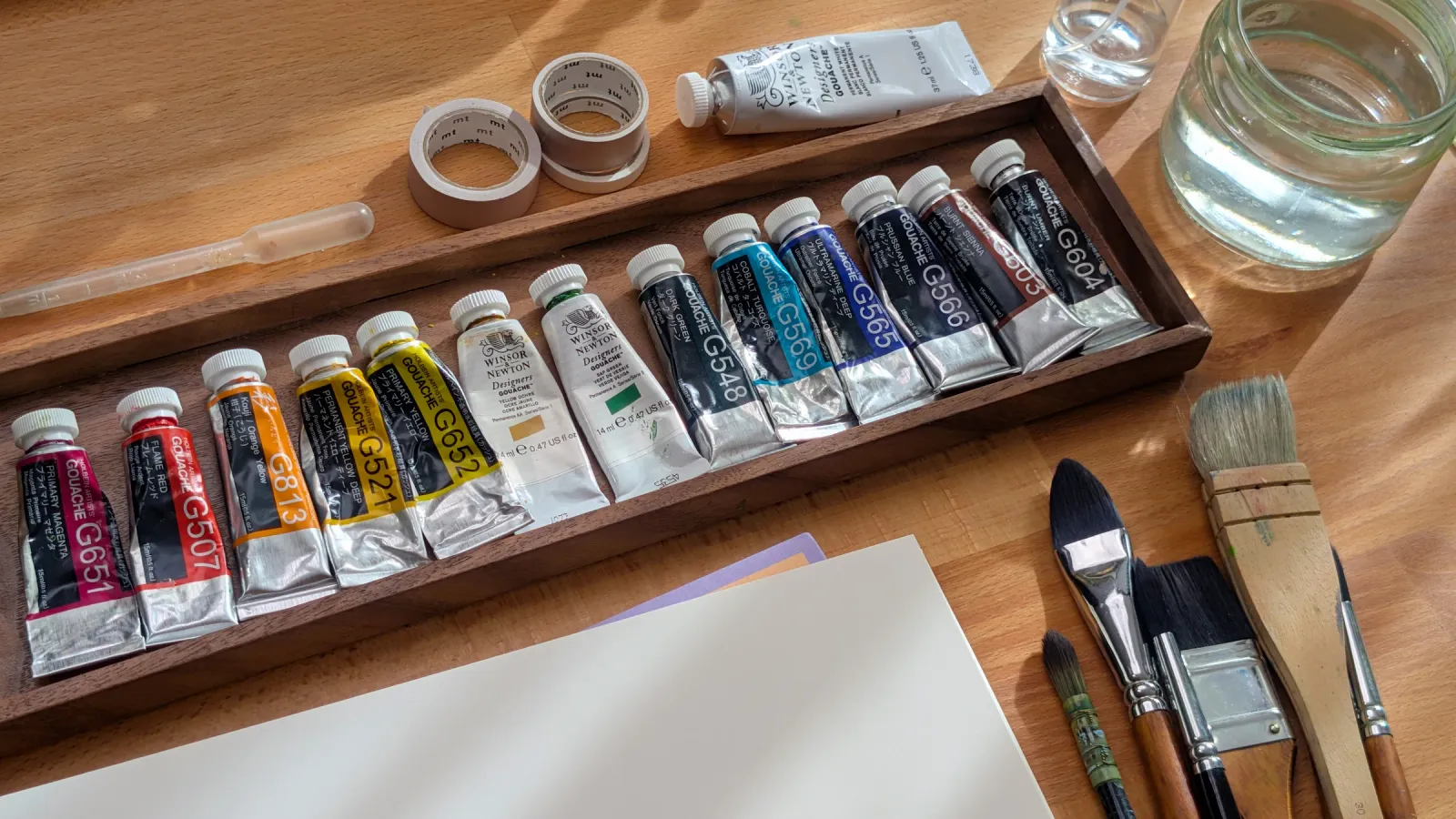 Gouache paint tubes lined up in a wooden tray on Maddy's desk